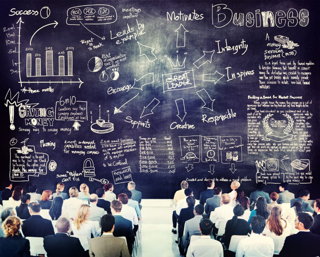 A large audience of business professionals, seen from behind, faces a giant chalkboard covered in hand-drawn business concepts, charts, and leadership terms.
